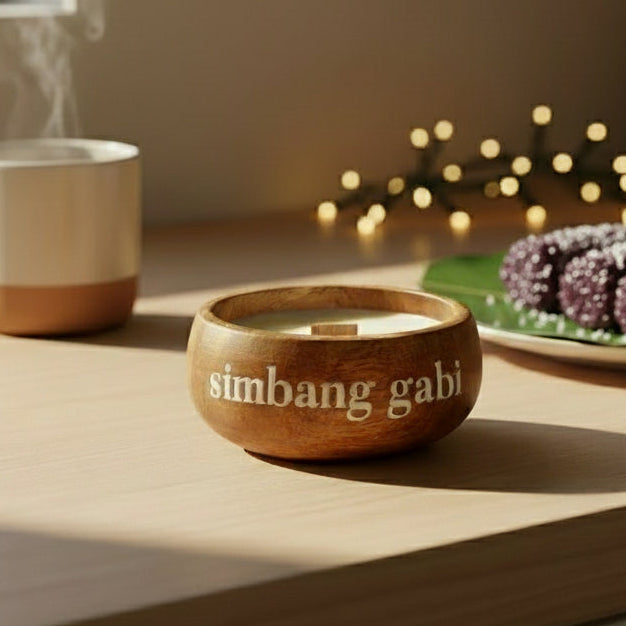 Wooden bowl labeled 'simbang gabri' with a lit candle, steaming mug, and plate of cookies on a kitchen counter.